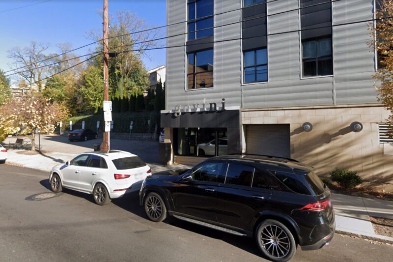Two SUVs are parked on a city street in front of a modern building with the sign "govini" visible on its facade. Trees and a sidewalk line the street.