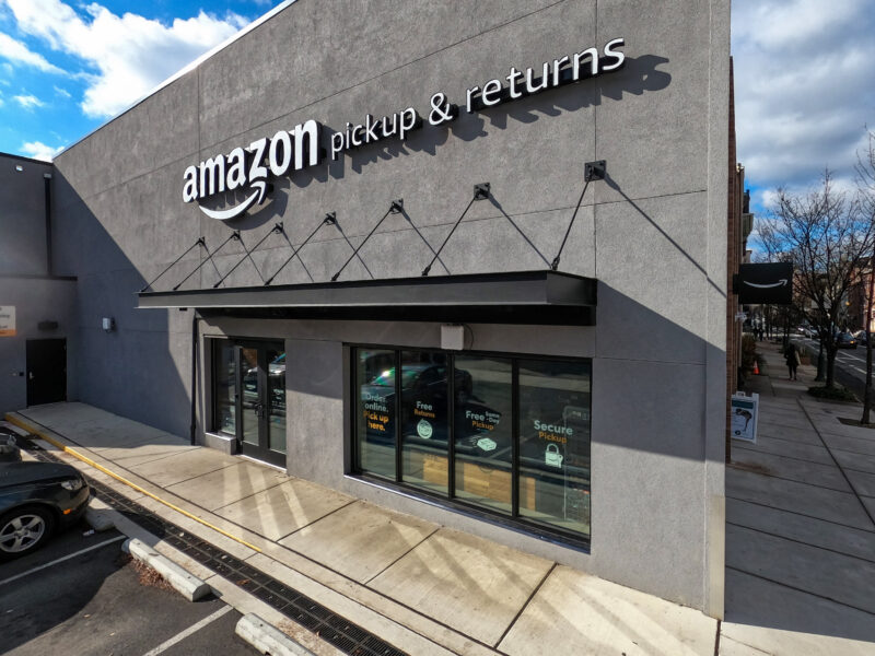 Amazon pickup & returns storefront with glass entrances, signs on doors, and a sidewalk in front; parked cars and trees visible nearby.