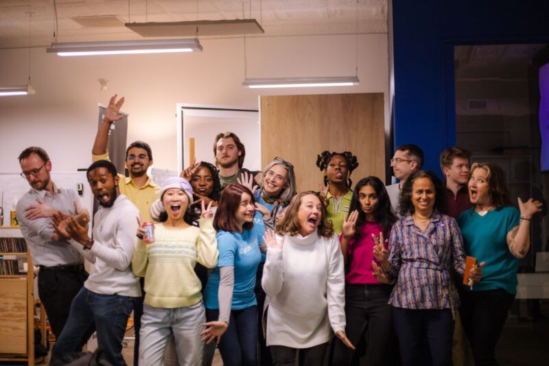 A group of people stand indoors, posing enthusiastically and making playful gestures, with some holding drinks.