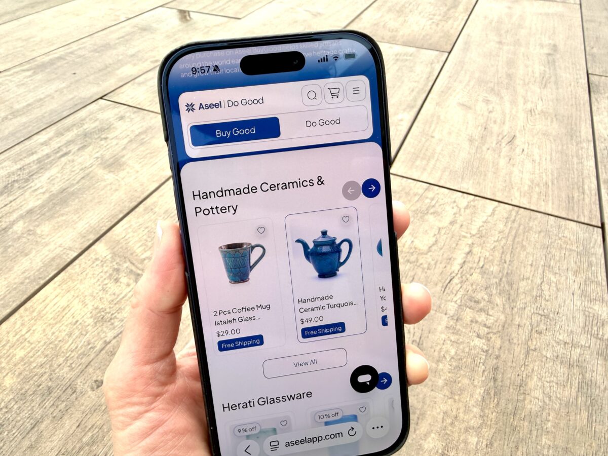 A hand holds a smartphone displaying a shopping app page for handmade ceramics and pottery, showing images and prices of items like mugs and teapots.