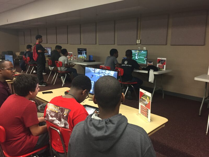 tudents sit in small groups at tables playing video games on monitors during an after-school esports event, with several others watching and waiting their turn.