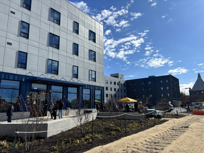A modern multi-story apartment building with people gathered near the entrance, adjacent landscaped areas, and a clear blue sky overhead.