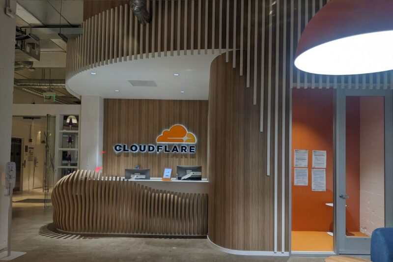 Reception desk at the Cloudflare office featuring a wood-paneled wall, company logo, and documents posted on an orange wall beside a glass door.