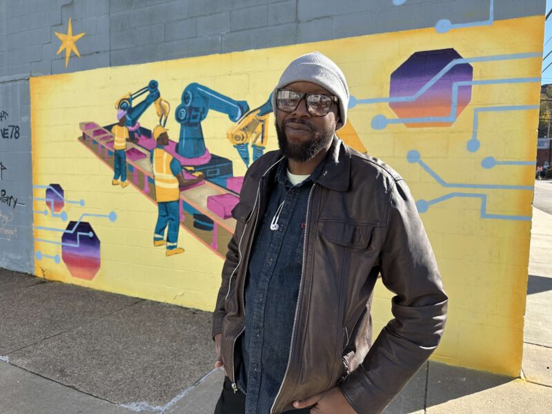 A man in a brown jacket and gray beanie stands in front of a colorful mural depicting industrial robots and people on a yellow background.
