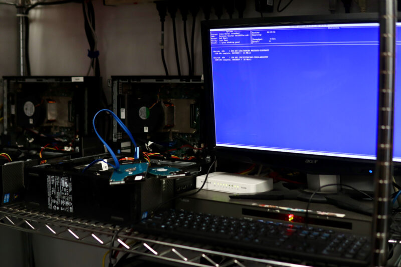 A computer monitor displays a blue BIOS setup screen next to several open desktop computer cases and hard drives on a metal shelf.