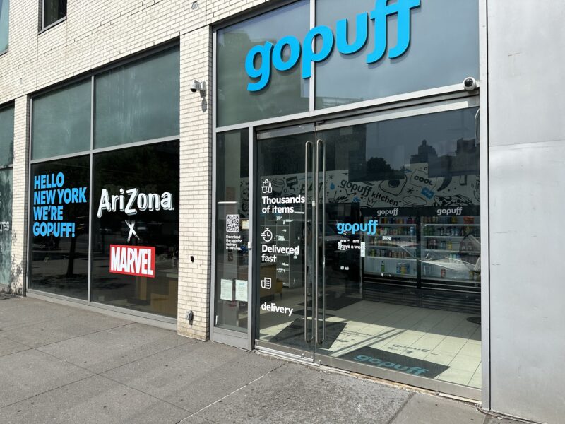 Street view of a GoPuff storefront with glass doors and windows featuring signs for GoPuff, AriZona x Marvel, and delivery service details.
