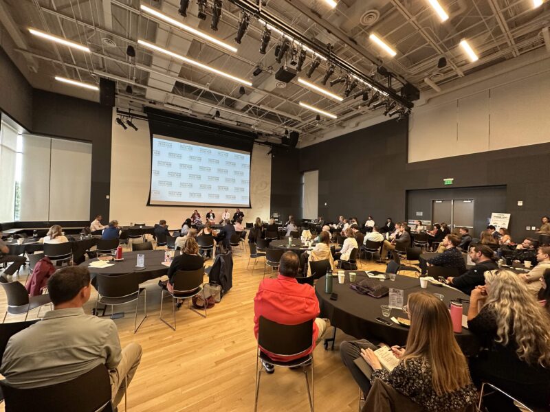 People seated at round tables listen to a panel discussion on stage in a modern conference room with a large projection screen.