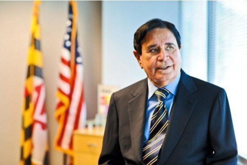 A man in a suit stands in an office with the Maryland state flag and a U.S. flag in the background.