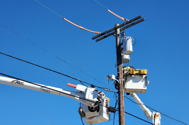 Two utility workers in bucket trucks repair electrical lines on a wooden utility pole under a clear blue sky.
