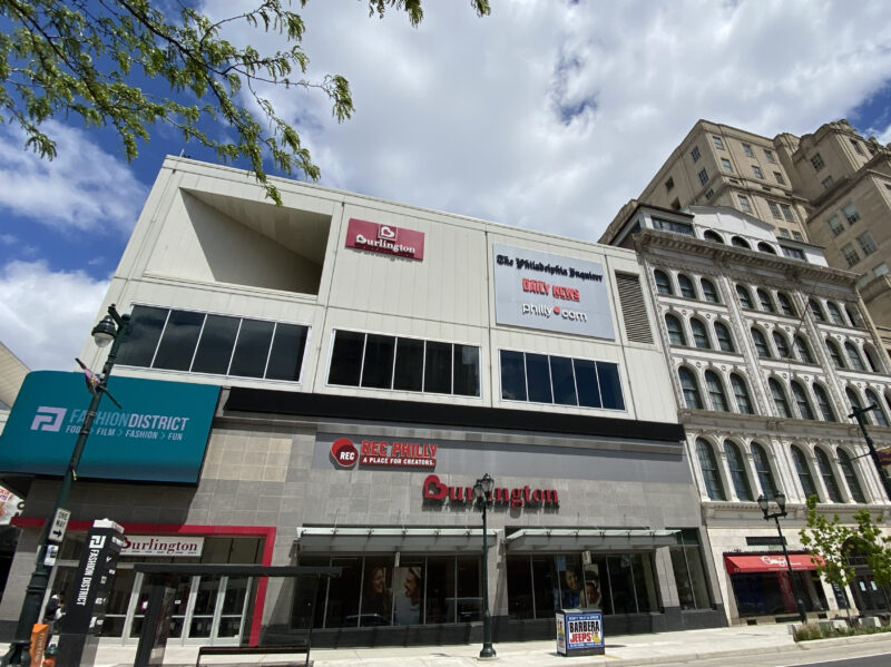 Street view of a modern retail building with Burlington and Fashion District signage, large windows, and an adjacent older multi-story building under a partly cloudy sky.