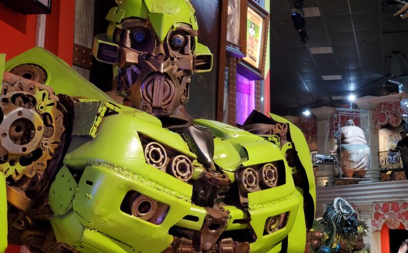A close-up of a green robot sculpture made from car parts is displayed indoors, with various decorations and statues in the background.
