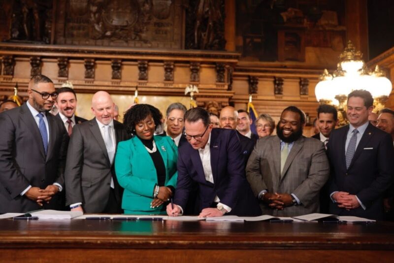A group of people in formal attire stand around a man signing a document at a table in a wood-paneled room with ornate decor.