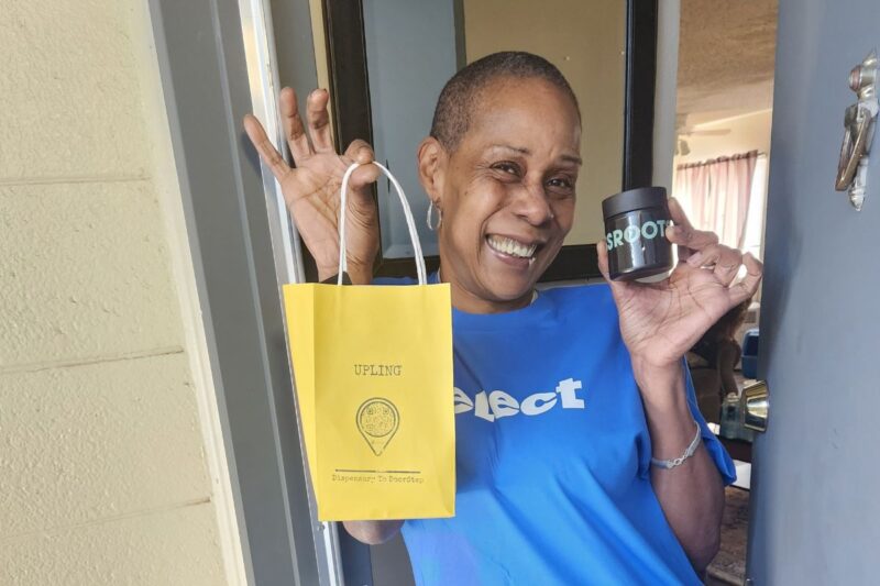 Smiling woman standing in a doorway holds a yellow UPLIFTN gift bag in one hand and a small black container labeled "SPROUT" in the other.