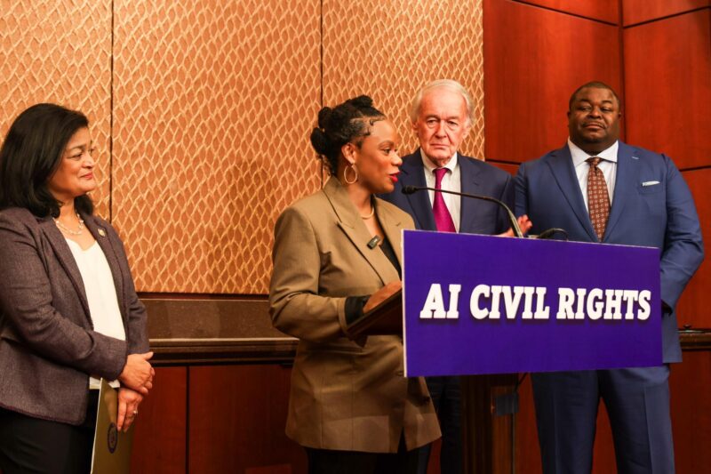 Four people stand at a podium labeled "AI Civil Rights"; one woman speaks while the others listen in a formal indoor setting.