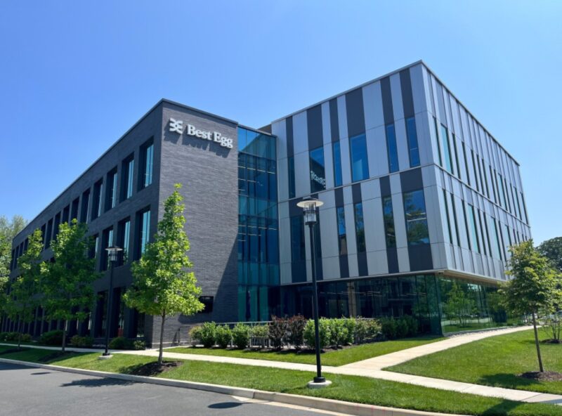 Modern three-story office building with "Best Egg" signage, large glass windows, and surrounding green landscaping under a clear blue sky.