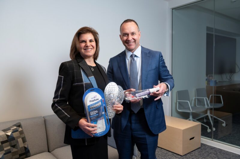 Two people in business attire stand indoors holding shoe insoles and related packaging, smiling at the camera. An office meeting room with chairs and a glass wall is visible in the background.