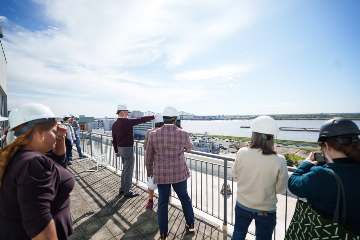 A group of people wearing hard hats stand on a balcony overlooking a river and a distant bridge under a clear sky.