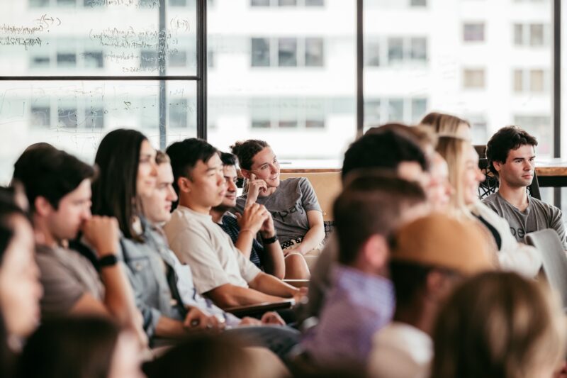 A group of people sitting closely together indoors, attentively listening to something out of view, with large windows in the background.