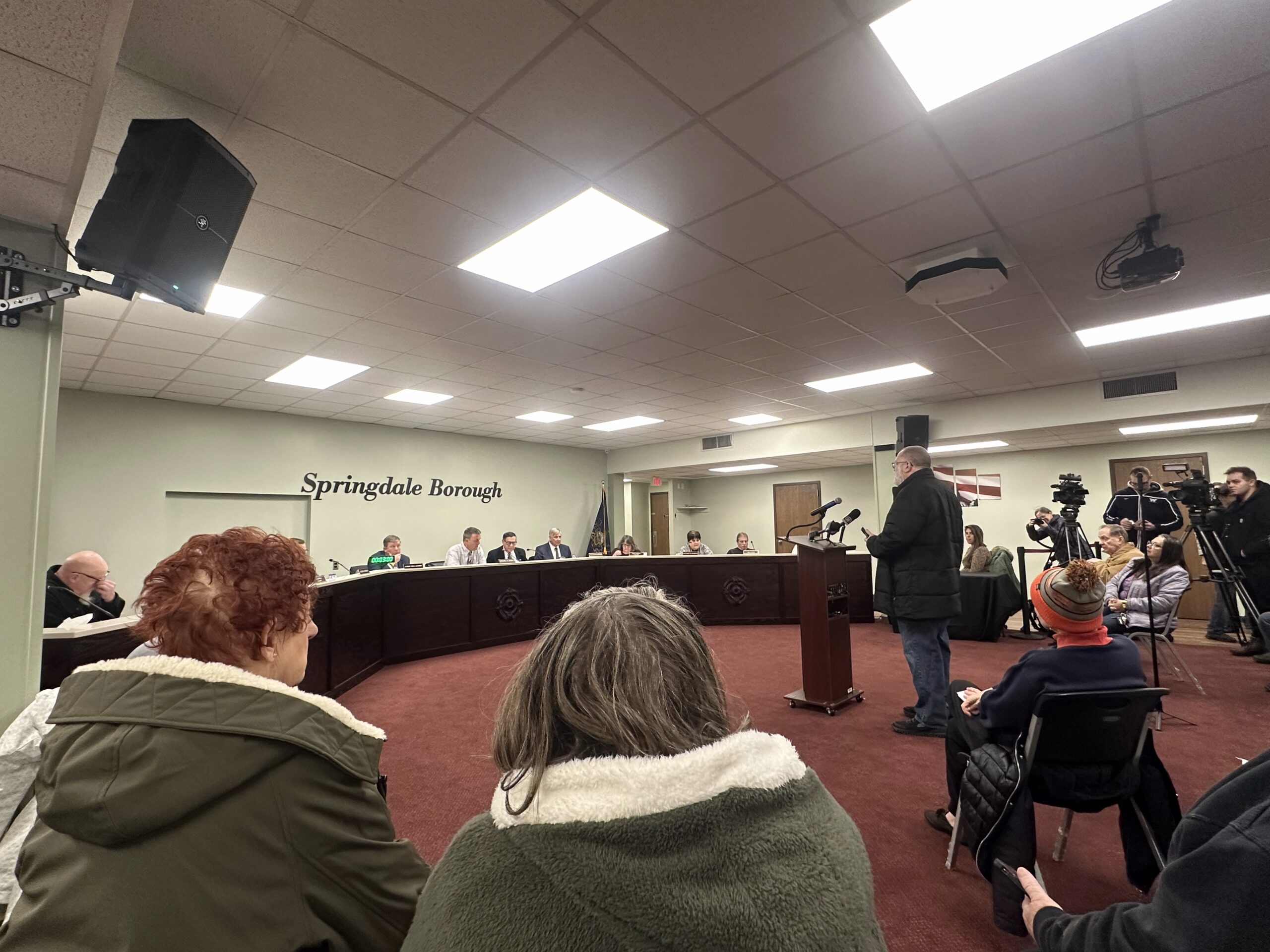 A man speaks at a podium during a public meeting in a room labeled "Springdale Borough," with council members, attendees and media present.