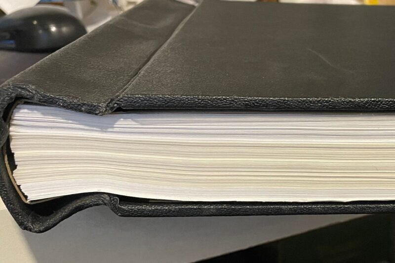 A close-up side view of a thick book with a black leather cover, showing many white pages stacked inside.