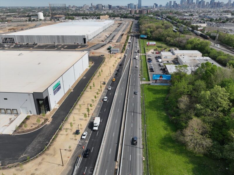 Aerial view of a multi-lane road with moderate traffic running between industrial buildings and green space, with a city skyline in the background.