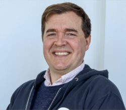 A man with short brown hair is smiling at the camera, wearing a dark hoodie with a circular logo and a collared shirt underneath, standing against a plain light background.