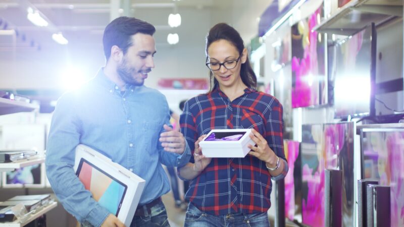 Two people walk through an electronics store aisle, holding boxes and looking at a tablet device together. TV screens are visible on the shelves beside them.