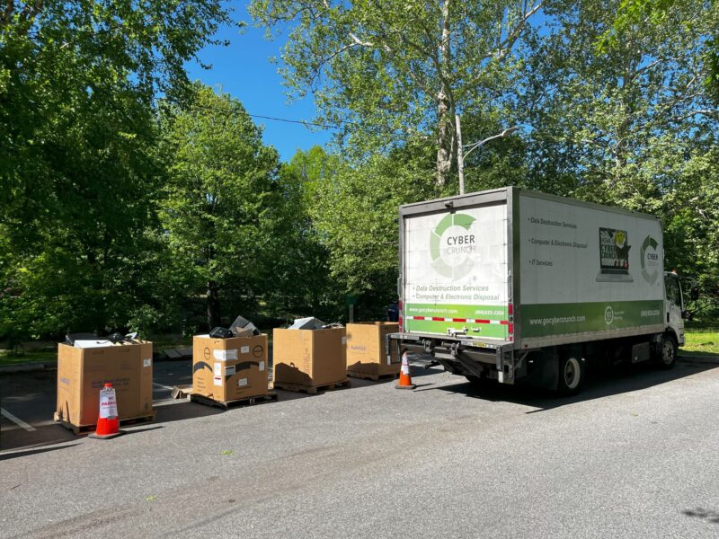A truck labeled "CyberCrunch" is parked next to large cardboard boxes filled with electronic waste in a tree-lined parking lot on a sunny day.