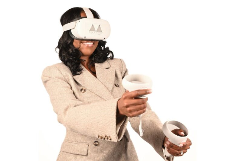 A woman wearing a beige blazer uses a virtual reality headset and holds two VR controllers against a plain white background.