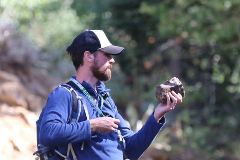 A man wearing a blue shirt, backpack, and a cap holds a large rock in one hand while standing outdoors in a wooded area.