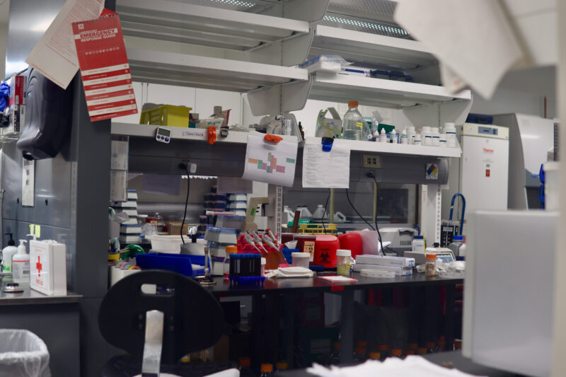 A cluttered laboratory workspace with various scientific equipment, bottles, containers, and papers scattered on benches and shelves.