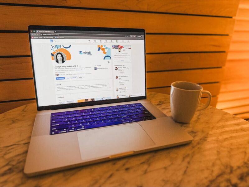 A laptop displaying a LinkedIn profile sits on a marble table next to a white coffee mug, with wooden paneling in the background.