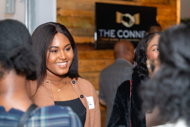 A woman smiles while engaging in conversation at an indoor networking event. A wooden wall and a sign reading "The Connect" are visible in the background.