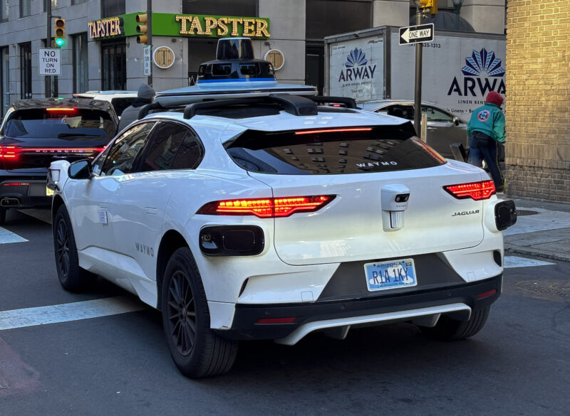 A white Jaguar SUV with Waymo branding and roof-mounted sensors is stopped at an intersection in an urban setting.