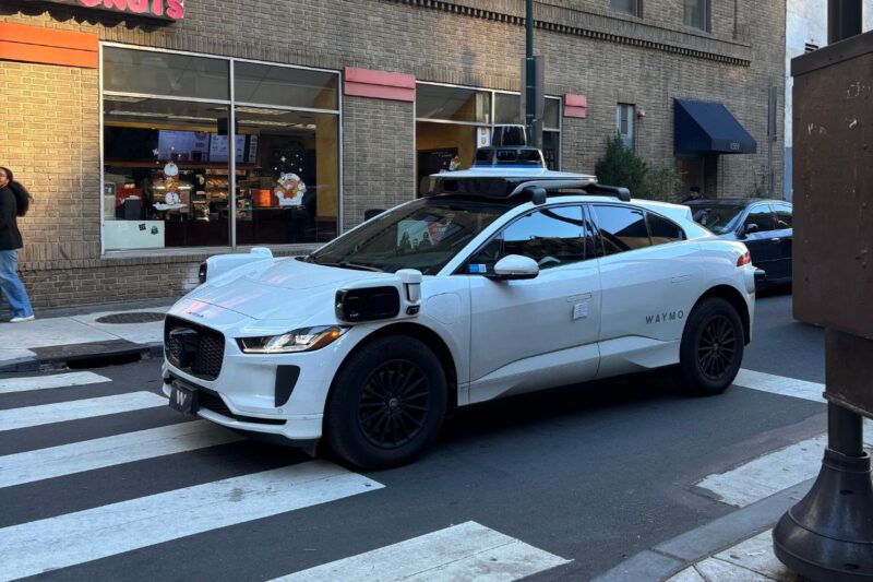 A white Waymo self-driving car is stopped at a crosswalk in an urban area, with storefronts and pedestrians in the background.