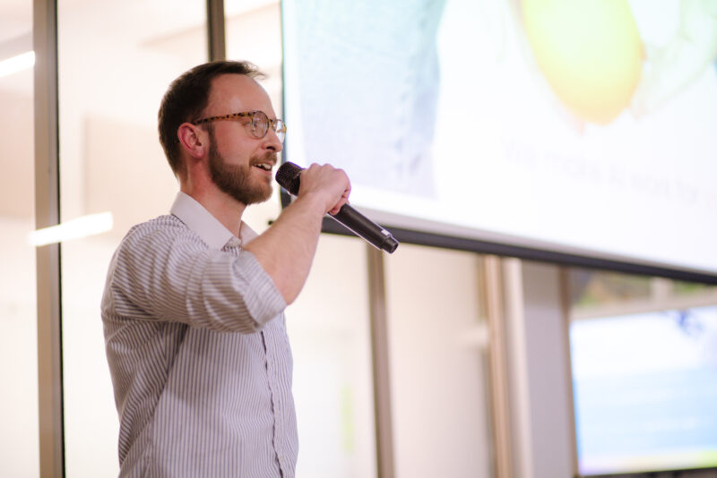 A man wearing glasses and a striped shirt speaks into a microphone in a brightly lit indoor setting, with a projection screen in the background.