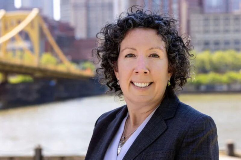 A person with short curly hair in a dark blazer stands outdoors near a river, with a yellow bridge and city buildings in the background.