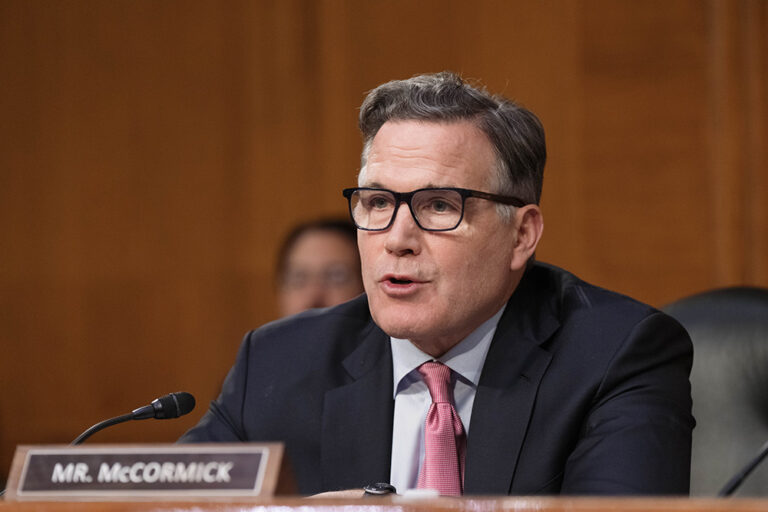 A man in a suit and glasses speaks at a microphone during a formal hearing, with a nameplate reading "Mr. McCormick" in front of him.