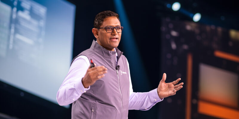 A man wearing glasses and a gray vest speaks on stage, gesturing with his hands, with large screens and stage lights in the background.