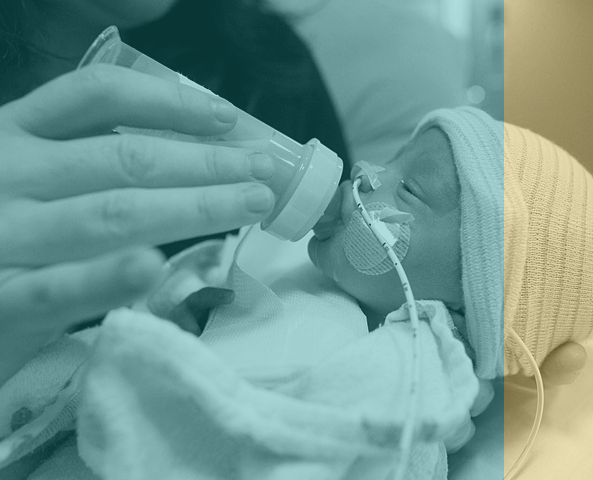 A newborn baby wearing a knit cap and medical tubes is fed from a bottle by an adult hand in a hospital setting.