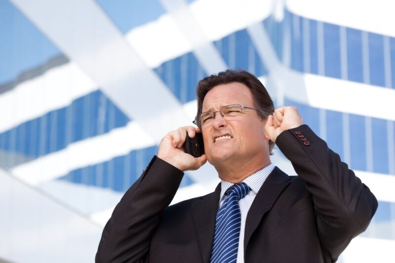 A man in a suit stands outside a modern glass building, appearing frustrated while talking on a mobile phone and covering one ear with his finger.