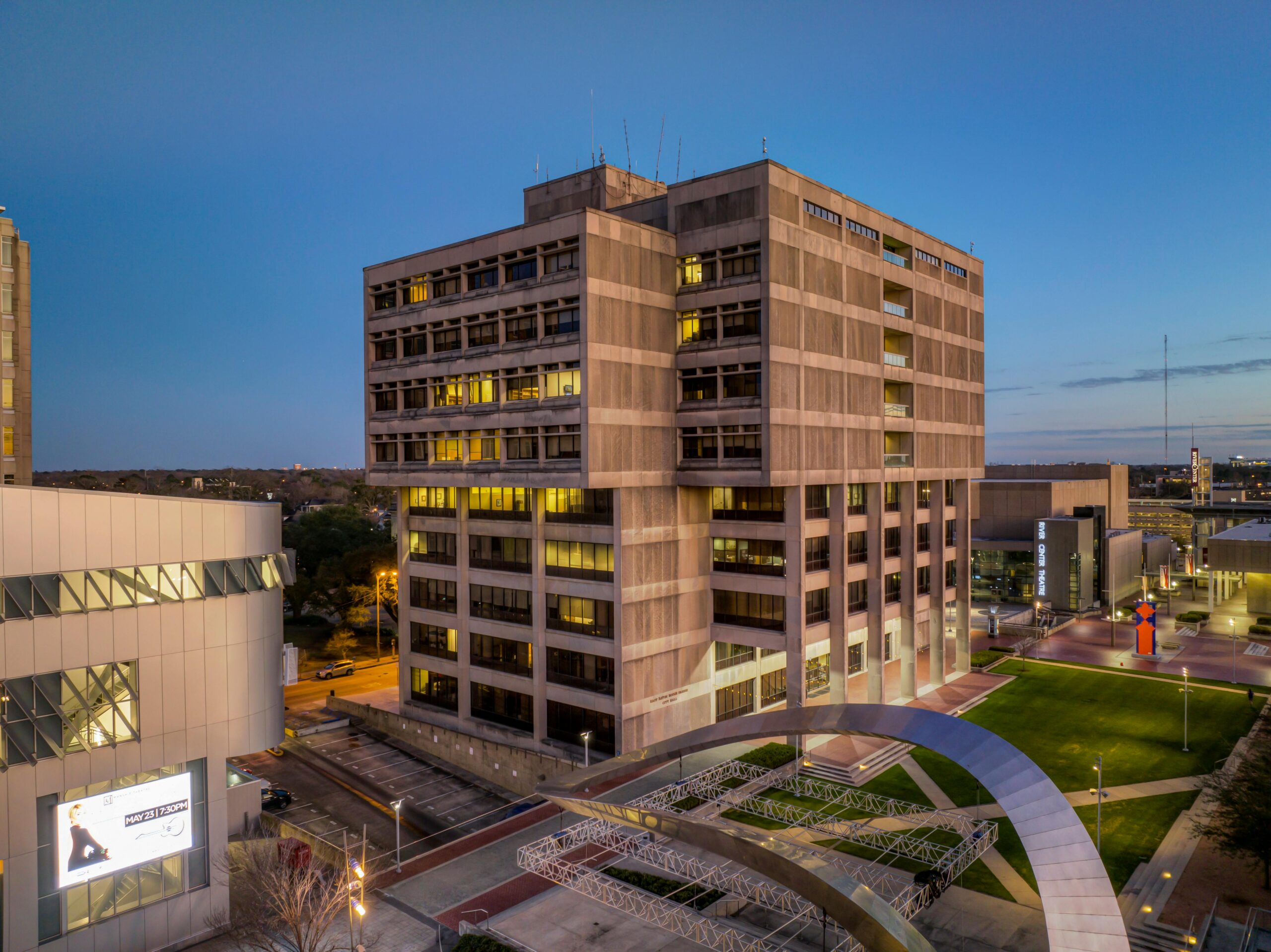 A large multi-story office building with exterior lights on at dusk, surrounded by modern structures and a landscaped plaza.
