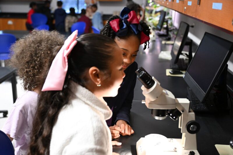 Three young girls observe a specimen through a microscope in a science classroom, with computers and cabinets visible in the background.