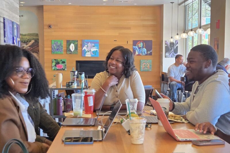 Three people sit at a table with laptops, drinks, and food, smiling and talking in a well-lit café with colorful artwork on the wall behind them.