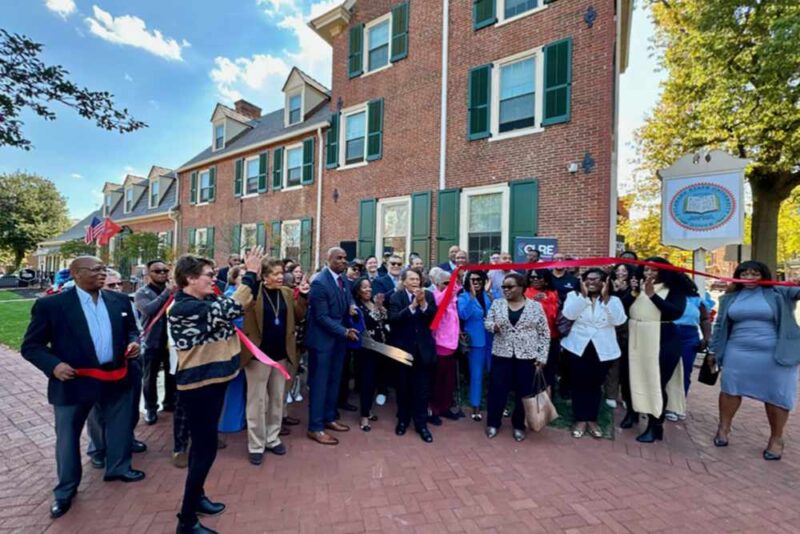 A group of people participates in a ribbon-cutting ceremony outside a brick building on a sunny day.