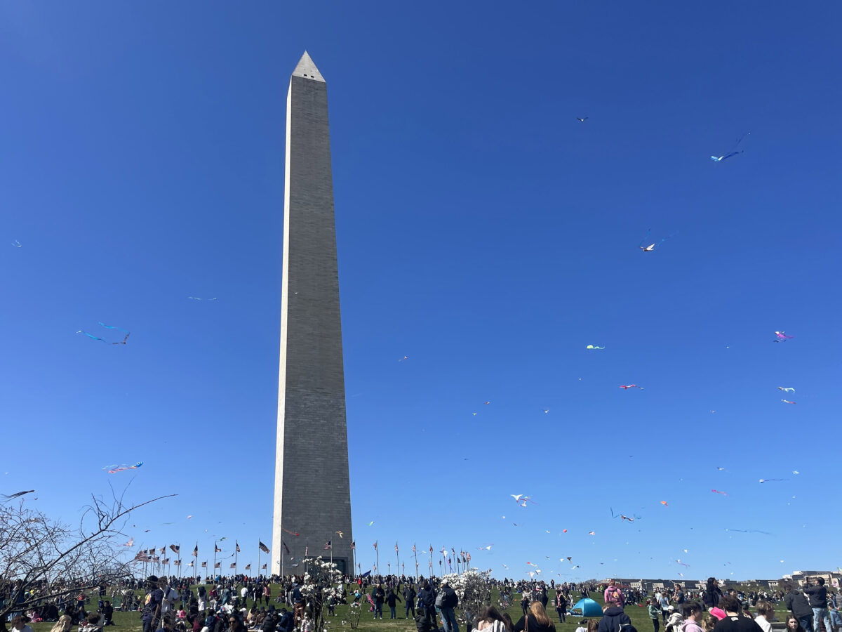 The Washington Monument with a crowd of people flying kites on a bright, clear day. Many kites are visible in the blue sky above the monument.
