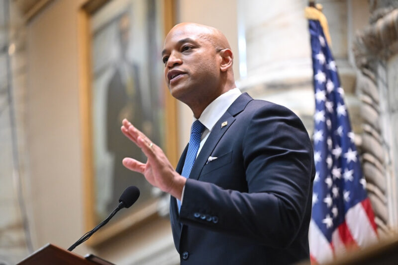A black man in a blue suit holds his hand up while speaking at a podium.