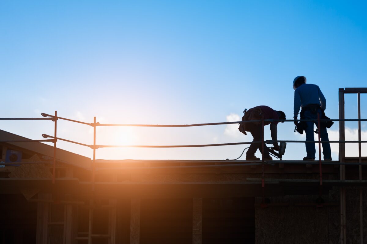 Two construction workers wearing safety gear work on a rooftop with a bright sky and sun in the background.
