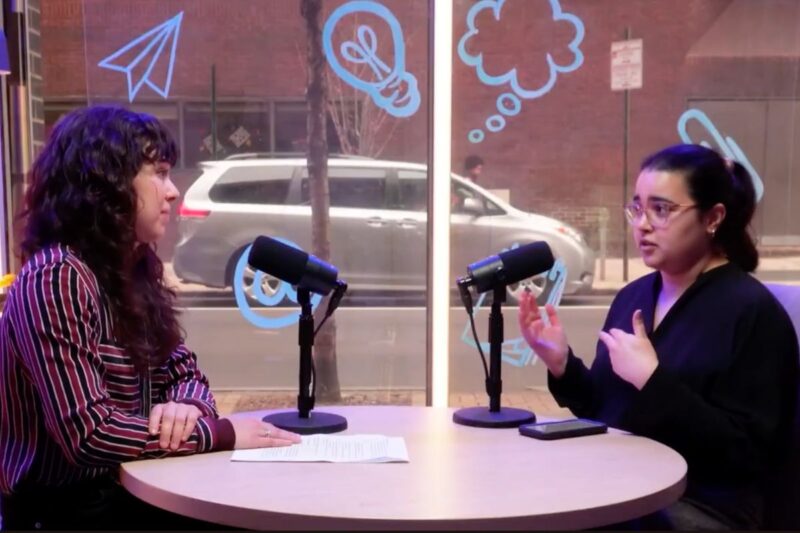 Two women sit across from each other at a round table with microphones, having a discussion. Blue illustrations decorate the window behind them, and a street scene is visible outside.