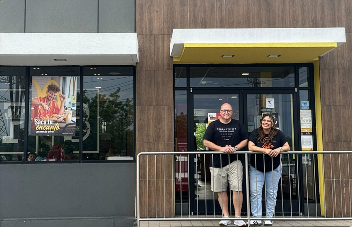 Two people stand smiling in front of a building entrance with a yellow awning, next to a window displaying a poster of a woman holding food.